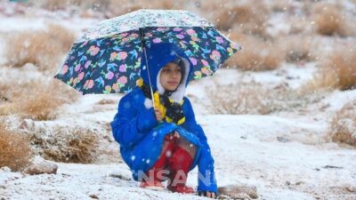 Tampak seorang anak kecil berbaju biru, dengan memakai payung serta baju tebal duduk bermain dengan salju di pegunungan Al-lawz, Tabuk, Arab Saudi.